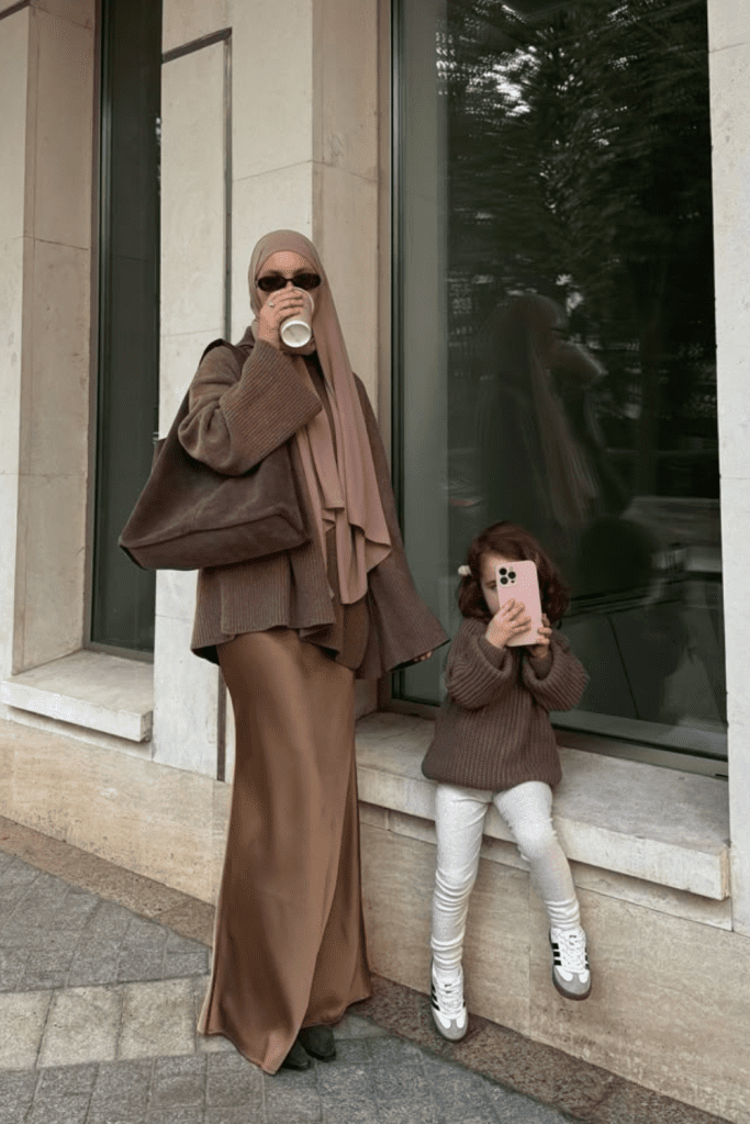 Hijabi mom in brown fall layers and her daughter in matching knit sweater posing together with warm coffee and smiles