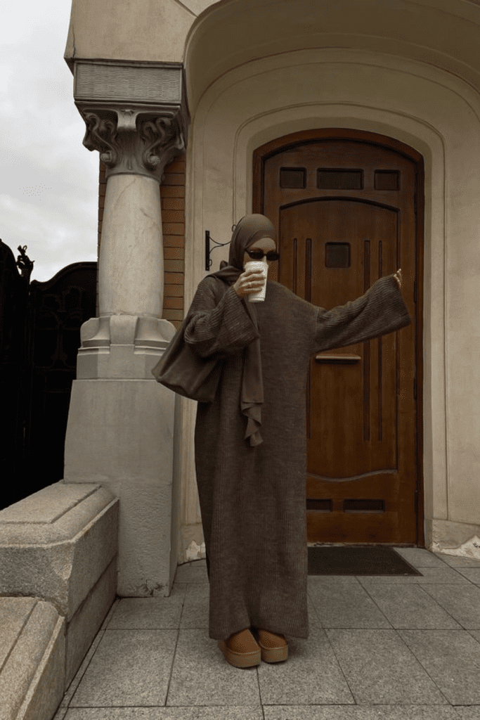 Muslim woman in a ribbed brown knit dress and hijab holding a coffee cup outside a wooden door in fall