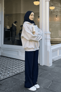 Muslim woman in a navy hijab and modest outfit with a light cream hoodie, wide-leg navy pants, and white sneakers. Holding a book outside a café or bookstore. Casual modest street style for fall.