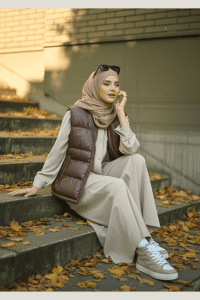 Young hijabi woman sitting on autumn steps wearing a beige shirt dress layered with a chocolate brown puffer vest, beige high-top sneakers, and a soft brown hijab. A warm and trendy modest outfit idea for early fall 2025.


