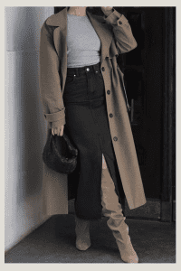 Casual fall outfit with a long camel trench coat layered over a grey top and high-waisted black denim skirt. Paired with over-the-knee taupe suede boots and a textured mini handbag. Chic modest city outfit.

