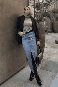 Hijabi woman in a fitted ribbed top, light denim maxi skirt with slit, black blazer, and black knee-high boots. Styled with a tan satin hijab and structured handbag. Modest fall streetwear with modern layering.

