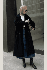 Hijabi woman styled in a black trench coat over a white shirt and long dark denim skirt, paired with a neutral satin hijab, sunglasses, heeled boots, and crossbody bag. Elegant modest fall outfit for city errands.