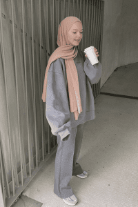 Hijabi woman wearing a grey oversized sweatshirt with matching wide-leg joggers, paired with a blush pink hijab and white sneakers, holding a coffee cup in an urban setting. Modest athleisure outfit for fall 2025.
