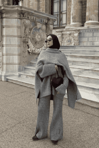 Hijabi woman wearing a ribbed knit co-ord set with wide-leg pants and an oversized sweater, styled with a matching shawl draped as a cape. Paired with sunglasses and a burgundy mini bag. Soft elegant layering for fall.
