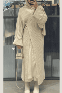Hijabi woman wearing a ribbed knit abaya-style cardigan layered over a white dress, paired with white ankle boots and a beige chiffon hijab. She carries a mini Jacquemus bag in a soft neutral shade. Cozy minimalist modest outfit for fall.