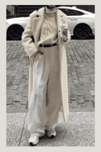 Elegant fall outfit with long cream coat, cable knit sweater, wide-leg trousers, and sneakers. Accessories include coffee cup, chain necklace, and black belt. Sophisticated modest fashion in monochrome tones.