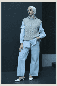 Hijabi woman wearing a pale grey cable knit sweater vest over a striped button-down shirt, paired with wide-leg light wash denim jeans and white pointed flats. Stylish and clean modest outfit for early fall.

