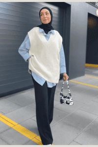 Hijabi woman in a white knit sweater vest layered over a powder blue button-down shirt, styled with black trousers, black hijab, and a mini cow print bag. Modest streetwear outfit idea for fall 2025.

